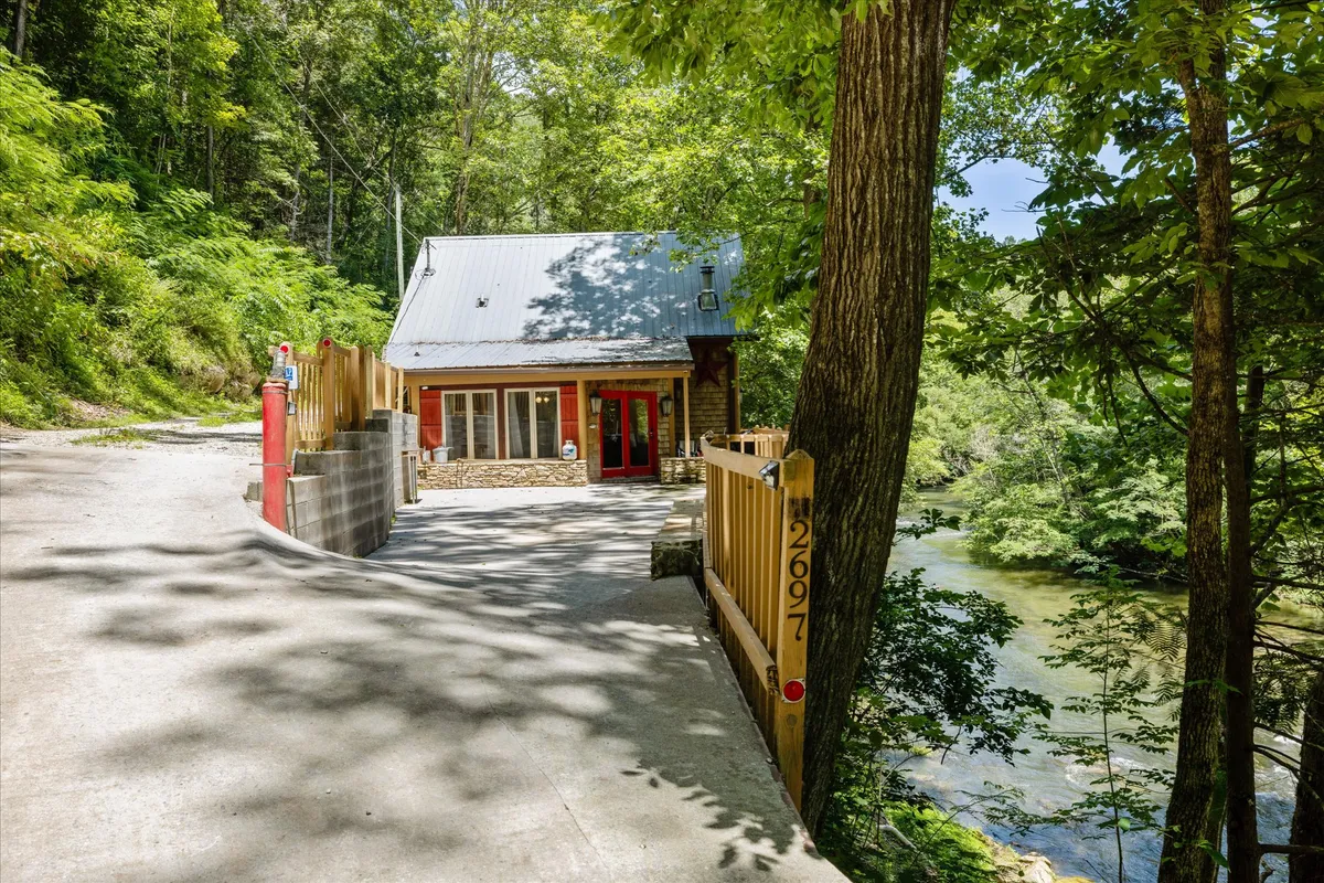 A wide exterior shot featuring the cabin with its inviting red door, nestled beside a gentle river and surrounded by lush trees. The setting sun casts a warm glow, highlighting the peaceful riverfront and expansive yard ideal for group gatherings.