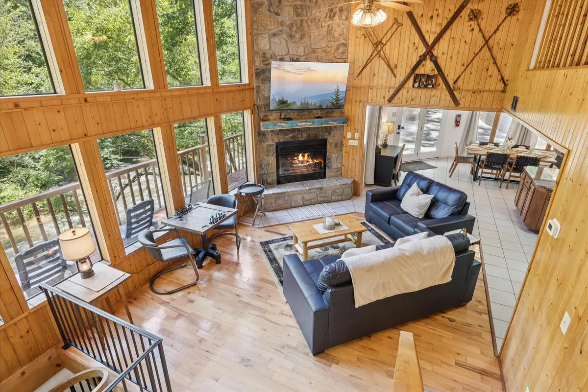 A spacious and inviting living room with large windows letting in natural light, a majestic stone fireplace at the center, comfortable seating arranged for conversation, and a chess table ready for games. The room feels warm, welcoming, and perfect for family gatherings.