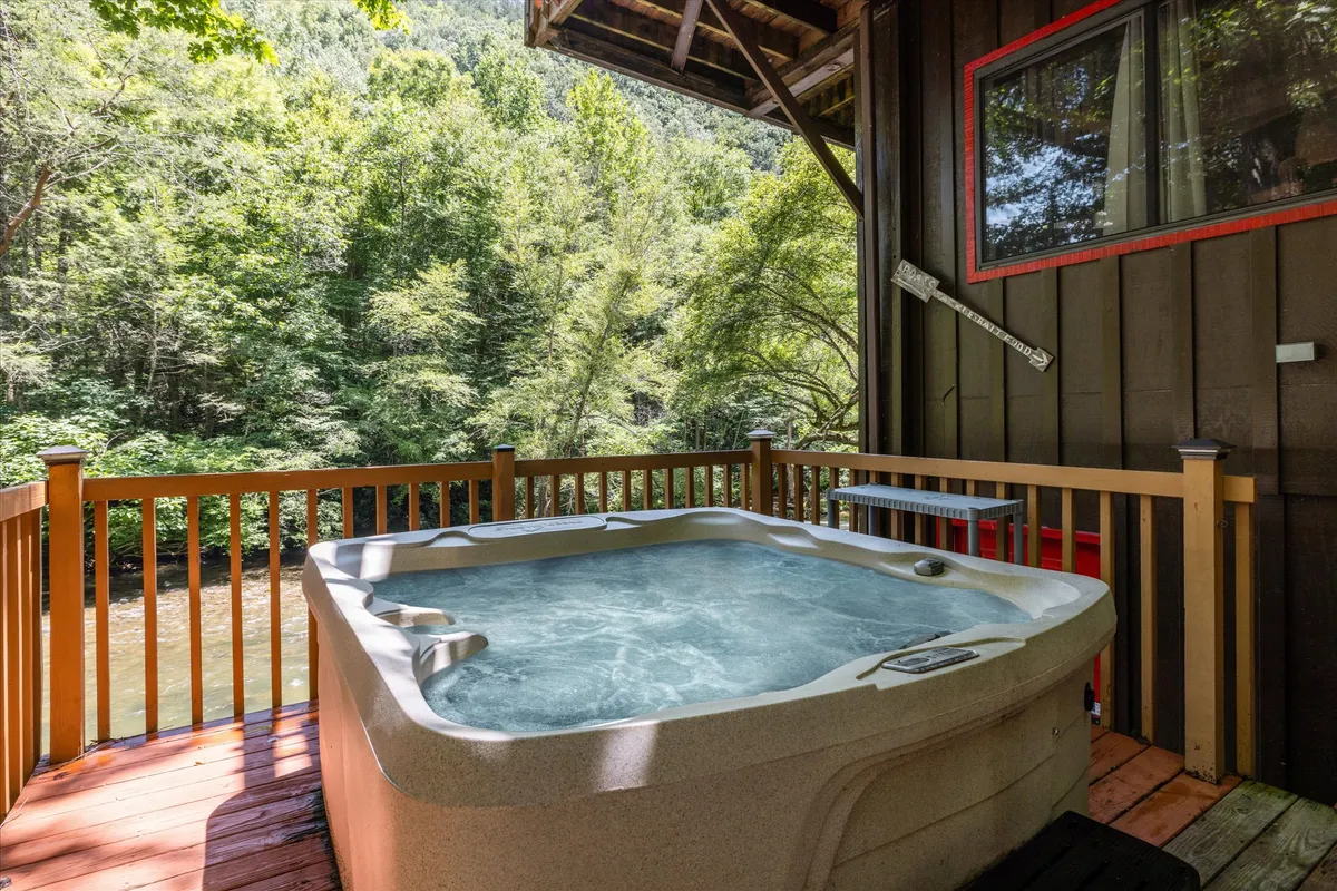 A tranquil hot tub set on a wooden deck surrounded by lush greenery. The hot tub overlooks the natural landscape, seamlessly blending the comforts of the cabin with the serenity of the outdoors. A portion of the wooden deck invites guests to relax and take in the river views, and the setting is ideal for stargazing or enjoying a morning coffee while immersed in nature.