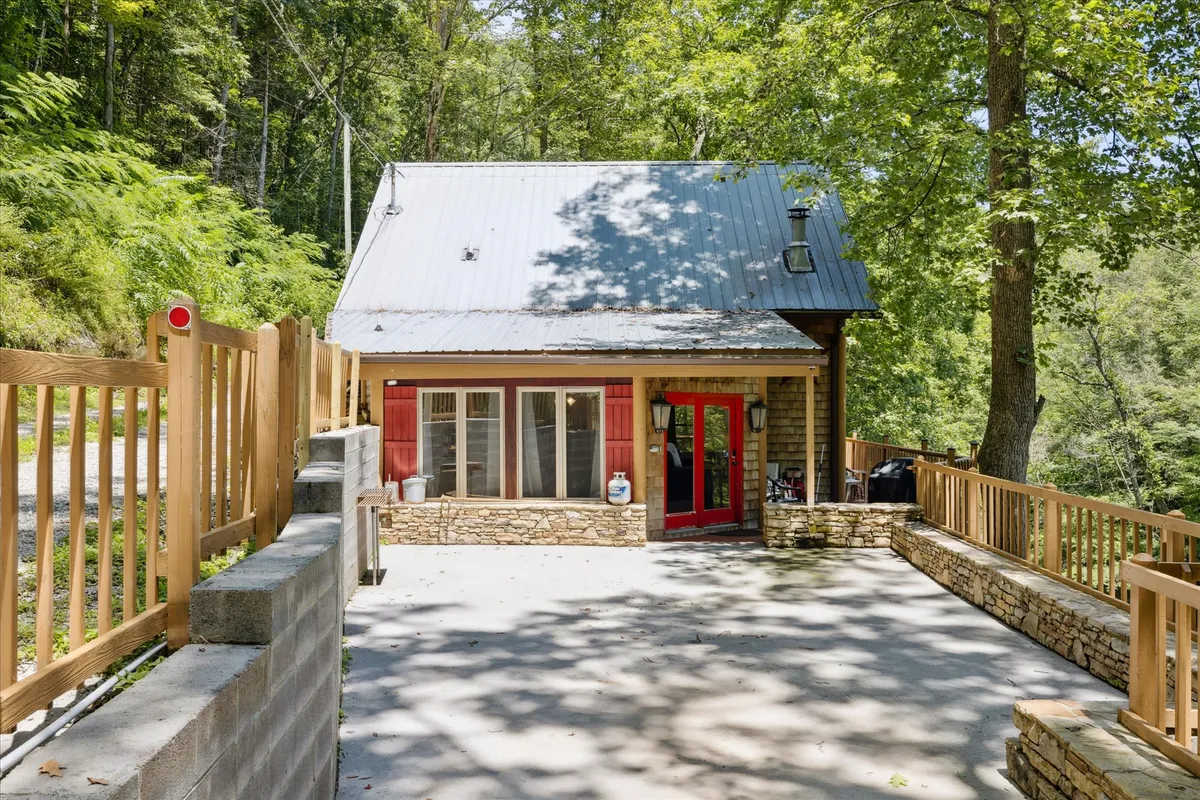 The image shows the welcoming exterior of a secluded riverfront cabin, featuring a charming red front door, rustic stone façade, and a wooden fence that hints at privacy. The patio invites morning coffee with nature views, while the spacious yard is perfect for relaxing and stargazing under Tennessee skies.