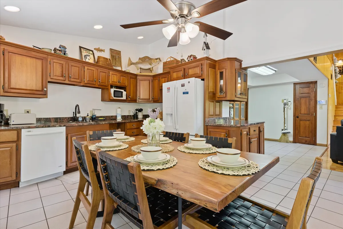 A cozy interior shot highlighting a wooden dining table set for a meal, surrounded by kitchen cabinetry. Dinnerware is neatly arranged, creating a scene set for family meals. Warm lighting and a ceiling fan overhead enhance the inviting atmosphere, and part of the kitchen is visible in the background, suggesting a seamless connection between the dining and kitchen areas.