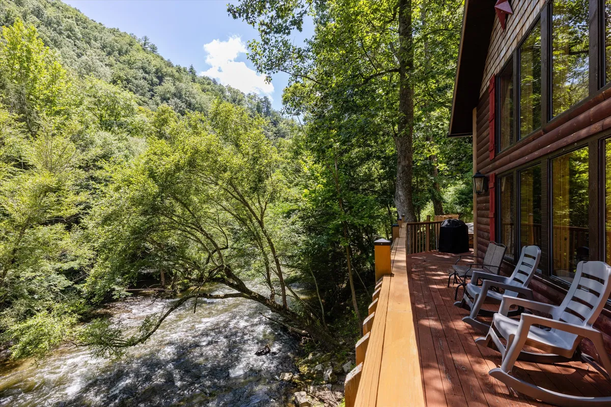 An exterior view from the cabin's deck overlooking the flowing river and distant mountains, surrounded by tall trees. Comfortable seating hints at relaxing moments, while the open space invites socializing or a barbecue as the sun rises or sets.