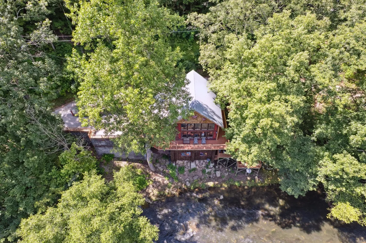A charming red cabin sits nestled by the river, with a cozy outdoor seating area surrounded by trees. The scene suggests peaceful mornings by the water, leisurely days spent outdoors, and evenings around a campfire or stargazing from the deck.