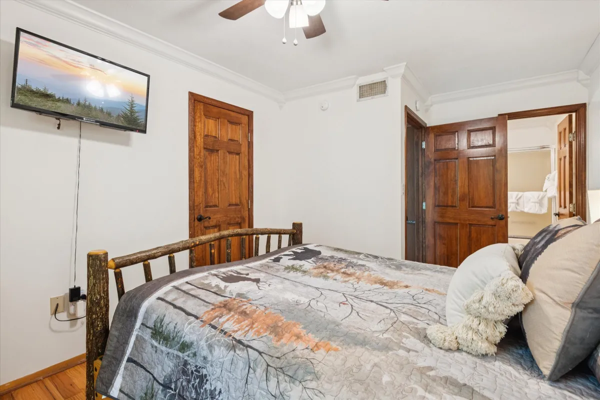 A comfortable bedroom with a wooden bed frame, decorative pillows, and a TV displaying a landscape scene. The cozy ambiance is designed for relaxation, with inviting textures and a view that suggests quiet, private downtime.