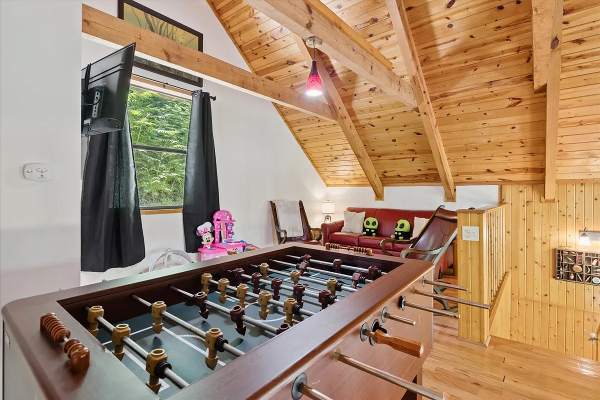 A bright, casual room featuring a foosball table, a comfortable sofa with decorative cushions, colorful plush toys, and wooden beams overhead. The setup invites playful activity and relaxation, with ample space for group games or lounging.