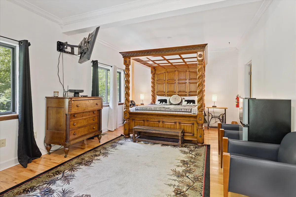 A serene bedroom with a classic four-poster bed, a wooden dresser, a floral-patterned rug, and windows that let in abundant natural light. The inviting setting is arranged for comfort and includes space for lounging with a book or enjoying morning coffee.