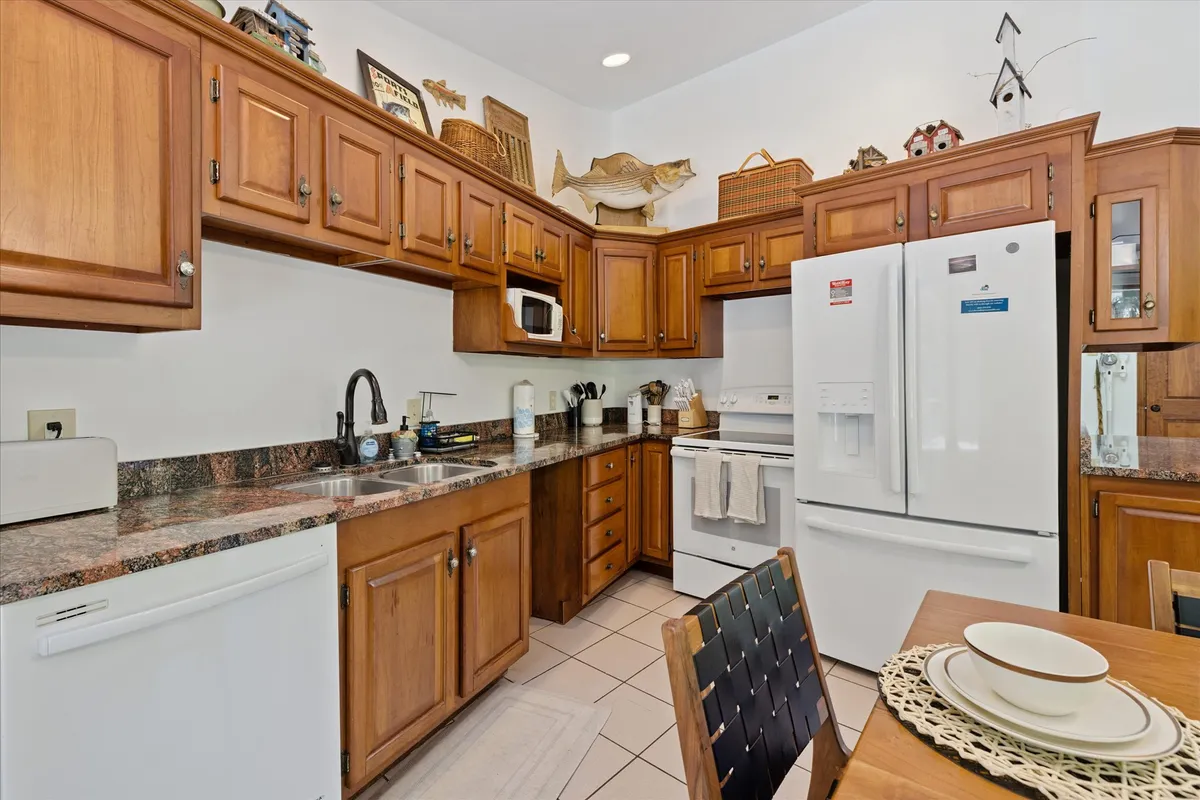 A view of the kitchen and adjoining dining table, featuring rich wooden cabinetry, granite countertops, and white modern appliances. The table is simply set, while natural light streams in, enhancing the warm and cozy feel. The open layout is visible, reinforcing the social, connected nature of the space.
